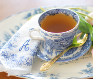A blue and white tea cup on a saucer. The cup is filled with tea. A gold spoon rests on the saucer. A white napkin with blue embroidery and the initial A is next to the tea cup.