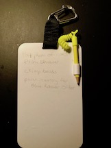 A small white plastic slate, suspended from a black fabric tab held by a silver carabiner. A yellow pencil is attached to the slate by means of a yellow cord. The slate has three notes written on it as example text.