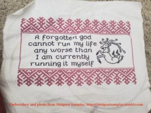 A white piece of fabric, embroidered with a red border. The center is stitched in black, with the words "A forgotten god cannot run my life any worse than I am currently running it myself". A deer-like creature in black thread is to the right of the words. A note at the bottom of the picture reads, "Embroidery and photo by Shitpost Sampler, https://shitpostsampler.tumblr.com"
