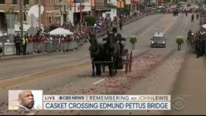 A screenshot of a black funeral carriage drawn by two black horses approaching the Edmund Pettus Bridge.