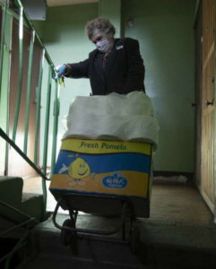 A woman wearing a face mask is looking at a note in her right hand, while she holds a hand truck with supplies at the top of a staircase in an unevenly lit hallway.