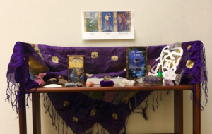 A triptych with images of Sigyn, Loki, and Angrboda on the wall, over an altar draped with a purple cloth with gold shapes and purple fringe, and adorned with various crystals, shells, and candles.