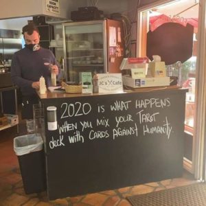A photo of a man standing behind the counter at a cafe. The chalkboard at the front of the counter reads, "2020 is what happens when you mix your Tarot deck with Cards Against Humanity".