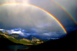 A photograph of a double rainbow, highlighted by puffy white clouds against a blue sky. The foreground is dark, and the background has green grass and two mountains.
