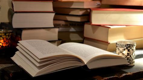 An open book sits between two glass candle jars. Stacks of books are arranged on the surface behind it.