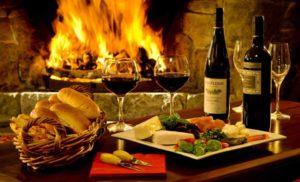 A table with a basket of bread, a plate of cheeses and fruits, two wineglasses filled with red wine, two bottles of wine, and two more empty glasses stans in front of a merrily burning fire in a stone fireplace.