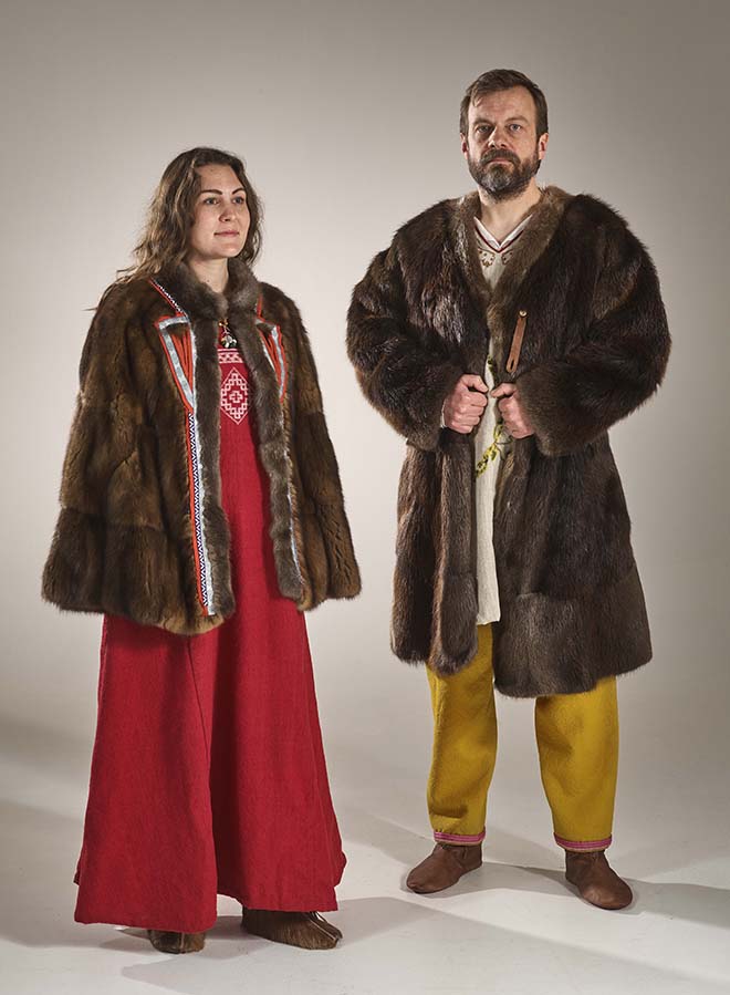 A medium-height woman with brown hair wearing a red linen dress with a white pattern woven into the bodice, leather slipper-style shoes, and a short fur cloak decorated in blue, red, and silver tablet-woven trim stands on the left of the photo. Next to her stands a taller man with short brown hair and a beard, wearing an undyed wool tunic with embroidery, dark yellow long pants edged at the ankles with red tablet-woven trim, and leather shoes. He wears a longer fur coat decorated with tablet-woven bands of wool, which hides most of the embroidery on his tunic.