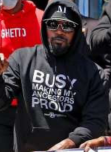 A photo of a Black man wearing a hoodie which reads "Busy Making My Ancestors Proud".