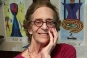 A photo of Rachel Pollack with her left hand resting against her face. She is standing in front of two of her paintings, and is beaming with joy.