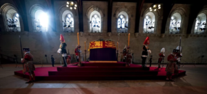A photo inside the main hall of the UK Parliament in London. Queen Elizabeth II lies in state. Her coffin is draped in the flag and surrounded by four tall candles on ornate stands. Members of the military stand guard.