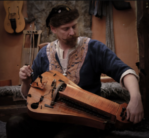A man in medieval Viking clothing is playing a hurdy gurdy.