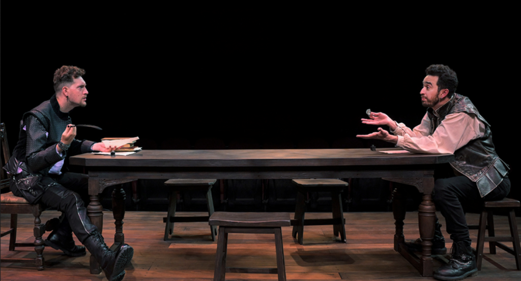 Two men, dressed as Christopher Marlowe and William Shakespeare, sit at opposite ends of a long table. They are gesturing emphatically as they discuss the play they are working on.